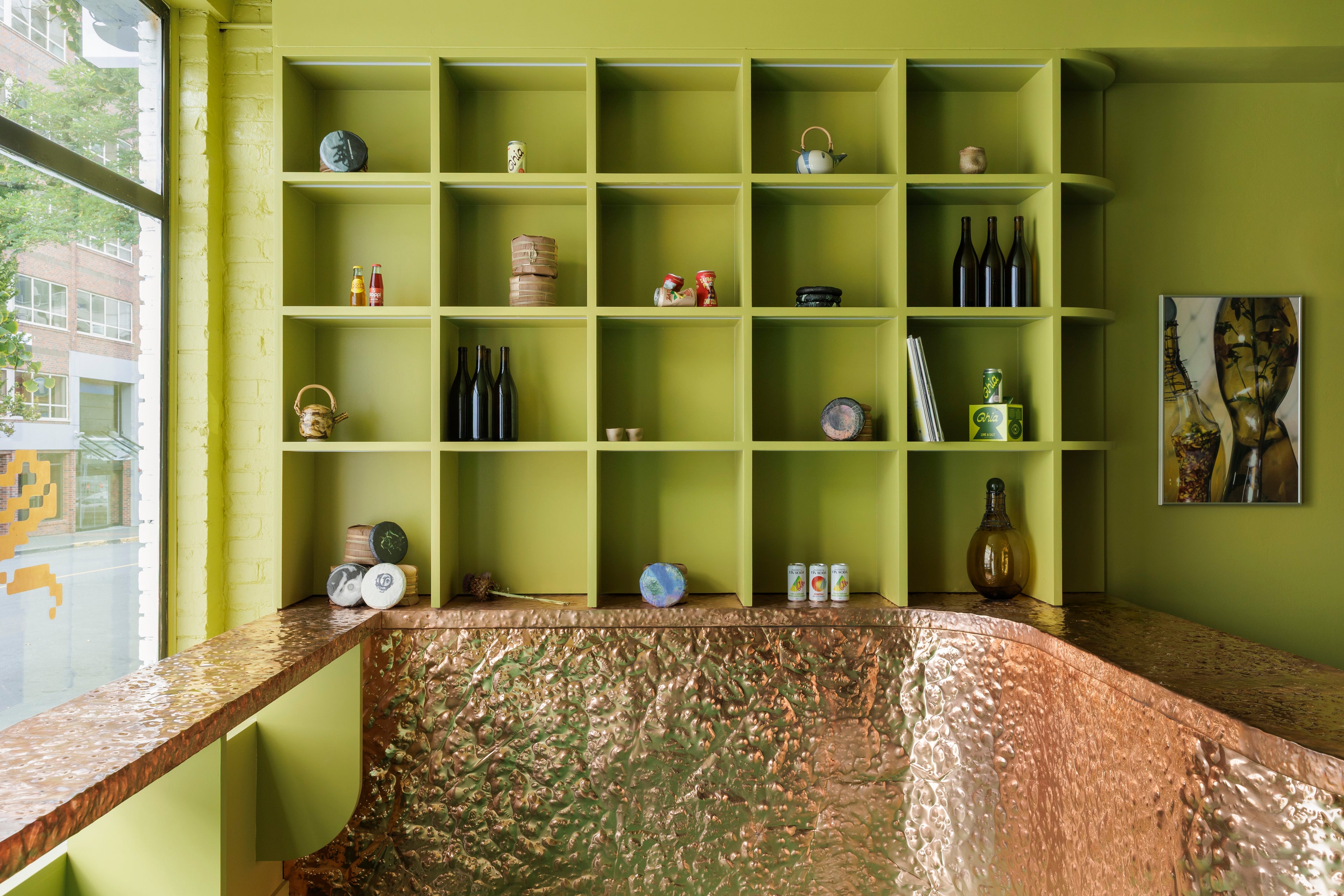 The interior of Softer Drink Shop, green built-in cabinets with various drinks, tea, and teaware above a hammered copper bar beside a large window.